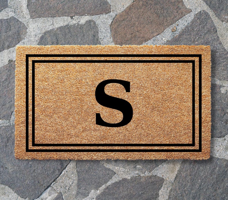 May include: A rectangular tan coir doormat featuring a bold black letter S centered within a double-lined black rectangular border. The natural fiber mat has a coarse, textured surface and a clean, geometric design printed in black ink.