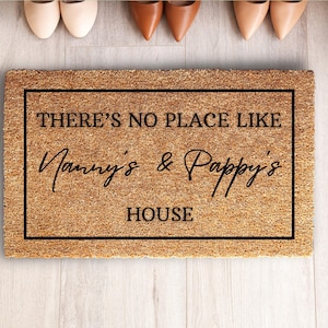 May include: A rectangular doormat made of natural fibers, featuring the text "THERE'S NO PLACE LIKE Nanny's & Pappy's HOUSE" in black script within a black border. The mat is placed on a light wood floor, with several pairs of shoes visible above.