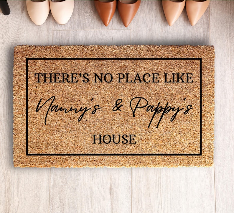 May include: A rectangular doormat made of natural fibers, featuring the text "THERE'S NO PLACE LIKE Nanny's & Pappy's HOUSE" in black script within a black border. The mat is placed on a light wood floor, with several pairs of shoes visible above.