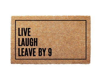 May include: A rectangular doormat made of natural coir fiber. The mat features a black border and the phrase "LIVE LAUGH LEAVE BY 9" in bold black letters, perfect for a home entrance.
