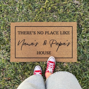 May include: A rectangular doormat with the text "THERE'S NO PLACE LIKE Nana's & Papa's HOUSE" in black lettering. The mat is brown and sits on green grass. Red sneakers are visible at the bottom of the image.