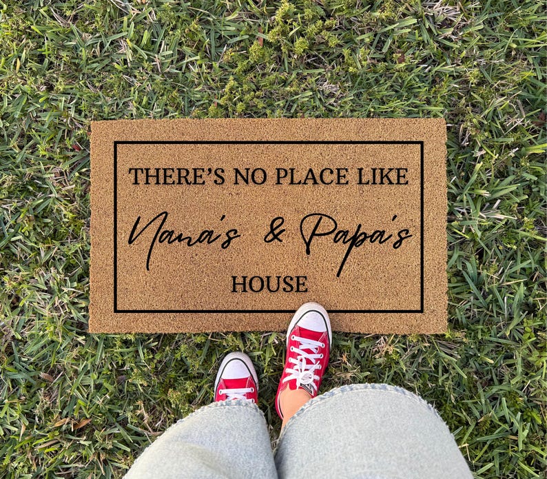 May include: A rectangular doormat with the text "THERE'S NO PLACE LIKE Nana's & Papa's HOUSE" in black lettering. The mat is brown and sits on green grass. Red sneakers are visible at the bottom of the image.