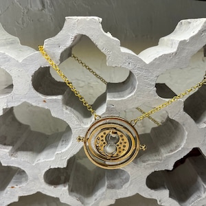 Può includere: Una collana dorata con un ciondolo rotondo che presenta un design a clessidra rotante. Il ciondolo è sospeso da una catena delicata.