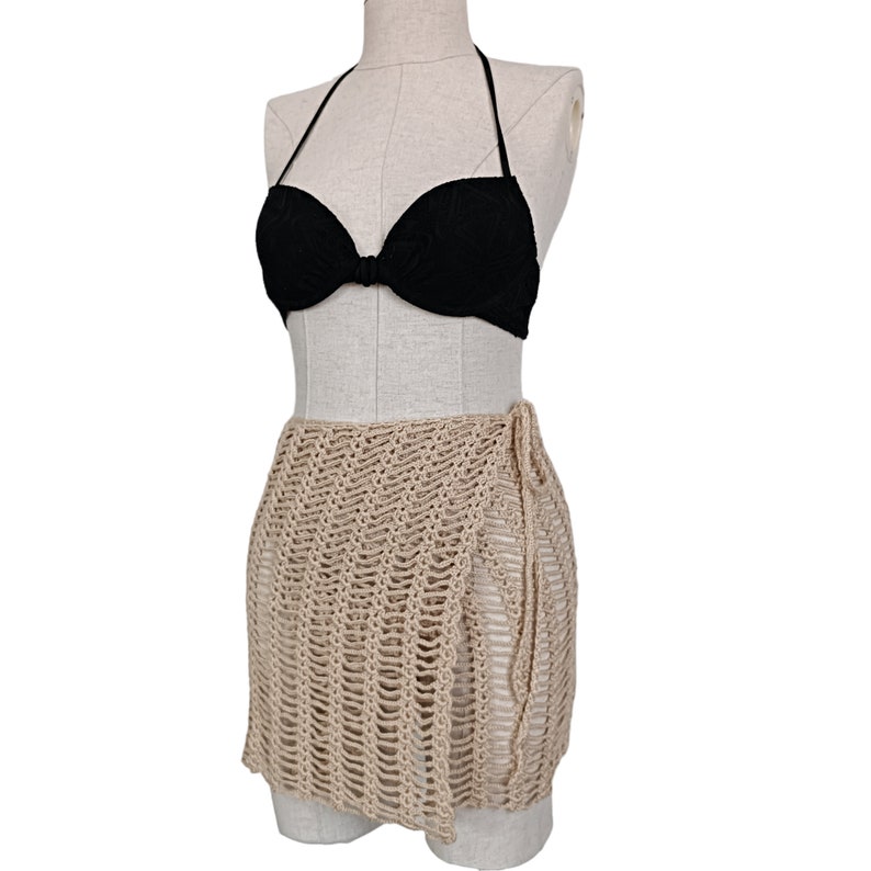 May include: A beige crochet wrap skirt with a tie closure. The skirt is made of a lightweight, airy fabric and is perfect for layering over a swimsuit.