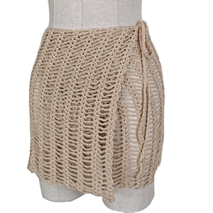 May include: A beige crochet wrap skirt with a tie closure. The skirt is made of a lightweight, airy fabric and is perfect for layering over swimwear or a sundress.