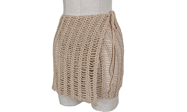 May include: A beige crochet wrap skirt with a tie closure. The skirt is made of a lightweight, airy fabric and is perfect for layering over swimwear or a sundress.