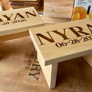 May include: Two light-colored wooden step stools. Each stool has a rectangular top and angled legs. One stool is engraved with the name "KAYAN" and the date "06-28-2026". The other stool is engraved with the name "NYRA" and the same date.