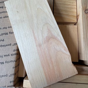 May include: A light-colored, rectangular piece of wood with visible grain and a slightly rough edge. The wood has a natural, unfinished appearance, with variations in color and texture. Other pieces of wood are visible in the background.