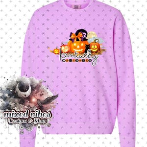 May include: A lavender sweatshirt with a graphic of pumpkins, a witch's hat, and a black cat. The text "Kentucky Halloween" is printed below the graphic. The sweatshirt has a crew neck and long sleeves.