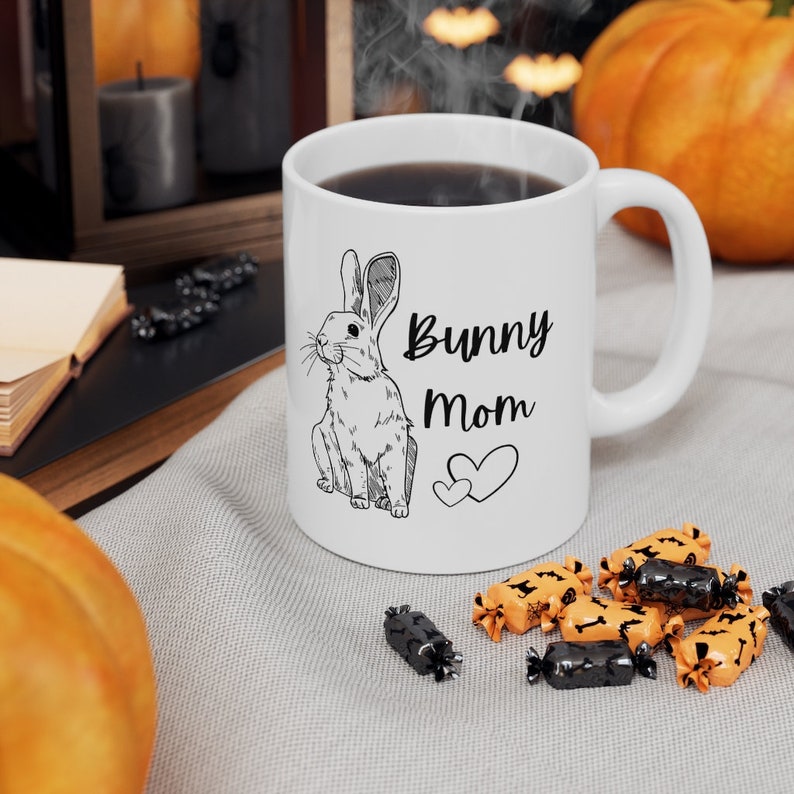 White ceramic mug with black design of rabbit and text that says "bunny mom" with two hearts.  Halloween themed decor surrounding the mug including candy and pumpkins.