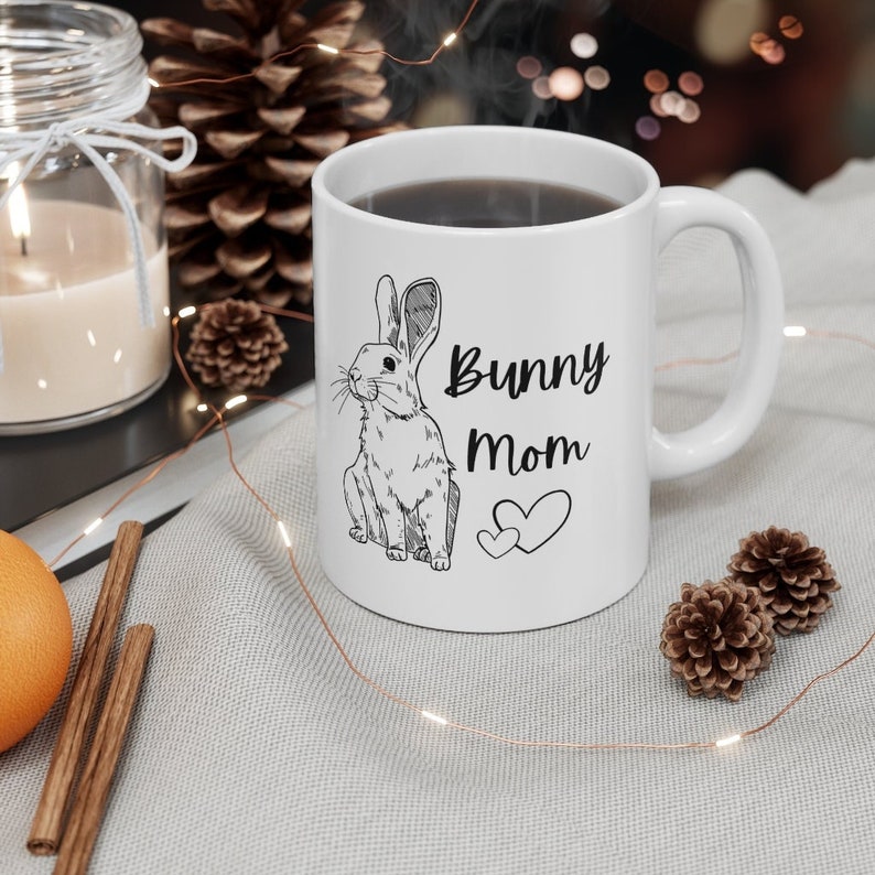 White ceramic mug with black design of rabbit and text that says "bunny mom" with two hearts. Mug is on a white tablecloth with fall theme decorations including a lit candle, pine cones, cinnamon sticks and LED gold wire light strand.