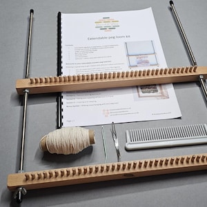 May include: An extendable peg loom kit featuring two light-coloured wooden beams with pegs, connected by long threaded metal rods with wing nuts. Includes a printed instruction manual, a spool of off-white yarn, two metal needles, and a white plastic comb.