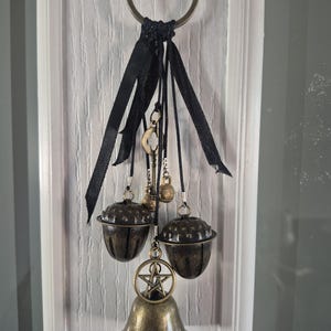 May include: A decorative hanging ornament featuring a large bronze-toned metal ring at the top. Attached are black ribbons and cords suspending two bronze acorn-shaped bells, a small skeleton key, a tiny bell, and a bronze pentagram charm above a bell.