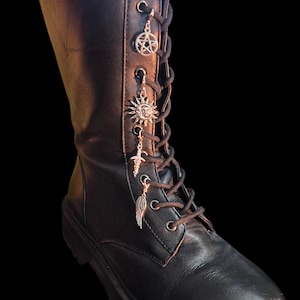 May include: Black leather combat boot with silver charms. Charms include a pentagram, sun, cross, and wing. The boot has black laces and a thick sole, set against a black background.