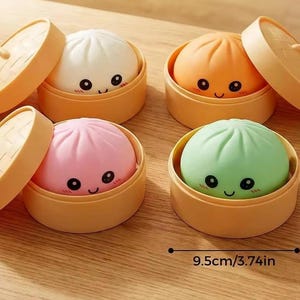 May include: Four round, smooth, colorful squishy toys shaped like steamed buns, each resting in a light brown plastic bamboo-style steamer basket with a matching lid. The buns are white, orange, pink, and green, each 7cm tall and 9.5cm wide.