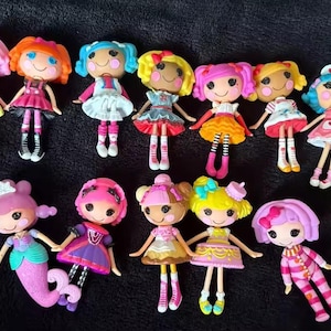 May include: A collection of colorful, small dolls with large eyes and rosy cheeks. Each doll has a unique hairstyle and outfit, including dresses, skirts, and accessories. The dolls are arranged on a dark surface.