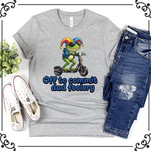 May include: A heather gray t-shirt featuring a cartoon frog riding a scooter, wearing sunglasses and a jester hat. The text "Off to commit dad foolery" is printed below the frog in blue. The shirt is paired with blue jeans and white sneakers.