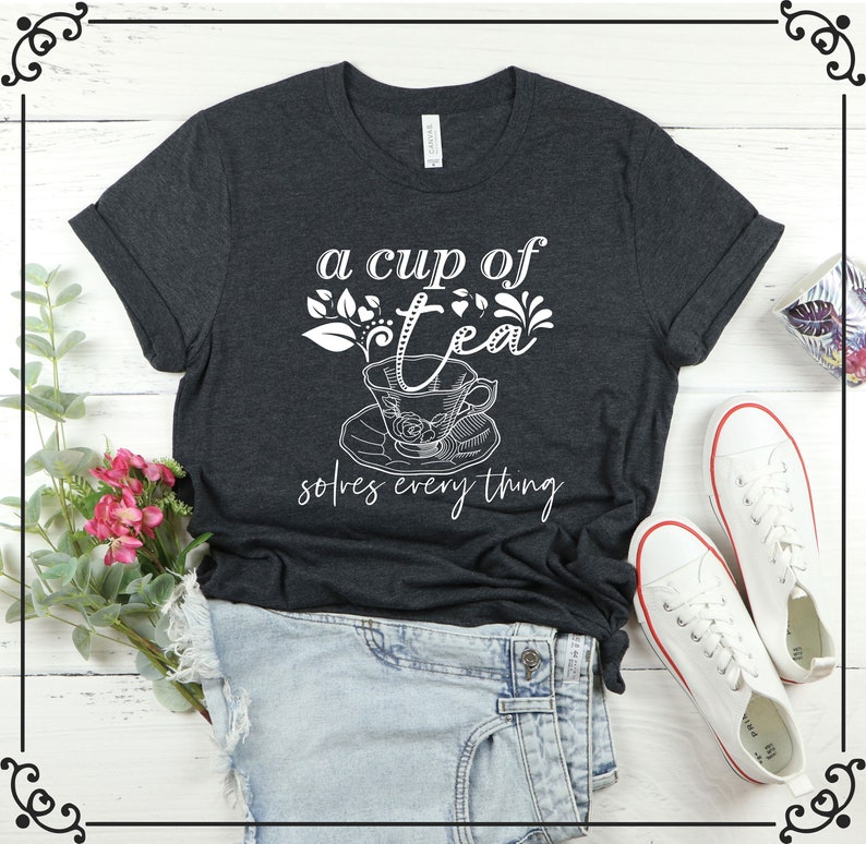 May include: Dark gray t-shirt with the text "a cup of tea solves everything" in white script. The design includes a teacup and floral accents. The shirt is paired with denim shorts and white sneakers.
