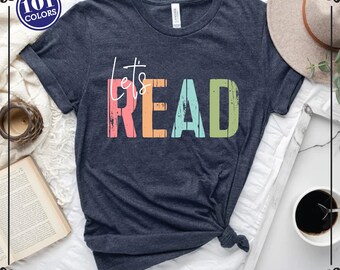 May include: A dark heather gray short-sleeve t-shirt featuring the white cursive text "let's" above the block-lettered word "READ" in distressed pink, orange, light blue, and green colors. A circular badge in the corner displays "101 COLORS".