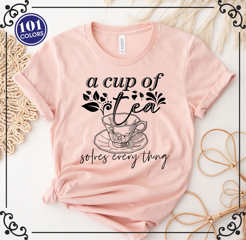 May include: A light pink t-shirt with the text "a cup of tea solves everything" in black script. The design includes a teacup and saucer illustration. A circular graphic in the upper left corner reads "101 COLORS".