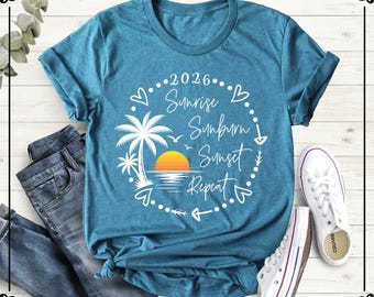 May include: A teal short-sleeve t-shirt featuring a white circular graphic with the text 2026, Sunrise, Sunburn, Sunset, Repeat. The design includes a palm tree, a yellow and orange sun, and small heart and arrow icons surrounding the text.