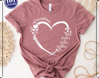 May include: A dusty rose t-shirt featuring a white heart design. The heart is adorned with butterflies and the word "grandma". Below the heart are the names Ryder, Luna, and Kohan. The t-shirt is knotted at the waist.