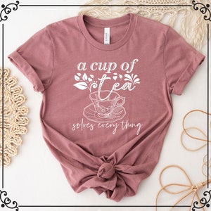 May include: Mauve t-shirt with the text "a cup of tea solves everything" in white script. The design includes a teacup and saucer with floral details. The shirt is knotted at the waist.