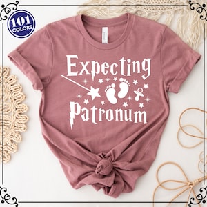 May include: A mauve colored t-shirt with the text "Expecting Patronum" in white. The text is accompanied by a white wand, a star, two white footprints, and a white pacifier.