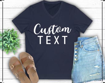 May include: A navy blue v-neck t-shirt with the text "Custom Text" in white. The shirt is photographed on a white wood background with a pair of brown sandals and blue denim shorts.