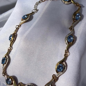 May include: A gold-toned necklace with oval, light blue gemstones and small, clear accent stones. The necklace has a delicate, flowing design with a mix of gold and silver-toned metal. The gemstones are evenly spaced along the necklace.