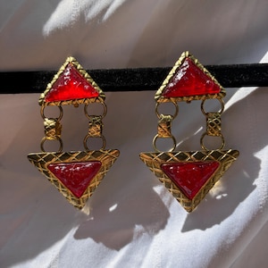 May include: A pair of gold-toned earrings with red triangular accents. The earrings feature a geometric design with a textured border around the red triangles and connecting links.