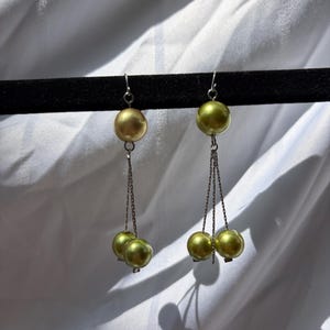 May include: A pair of dangle earrings with a gold and green color scheme. Each earring features a large, round gold bead at the top, connected by a chain to smaller green beads. The earrings hang from silver-colored hooks.