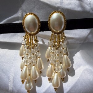 May include: A pair of vintage dangle earrings. Each earring features a large, oval, off-white faux pearl center, surrounded by a gold-toned frame. Below the center, strands of clear beads and teardrop-shaped faux pearls cascade downward, creating a glamorous, eye-catching design.