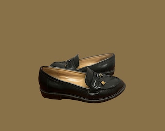 May include: A pair of black leather loafers with a gold-colored accent on the vamp. The shoes have a classic design with a rounded toe and a low heel. The loafers are displayed on a brown background.