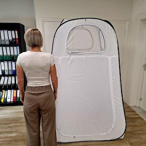 Può includere: Una tenda per la privacy bianca a comparsa con finiture nere e una finestra. La tenda è in piedi su un pavimento di legno. Una persona è in piedi di fronte alla tenda.
