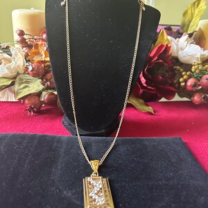 May include: A gold chain necklace with a rectangular pendant featuring a geometric design with clear crystals.