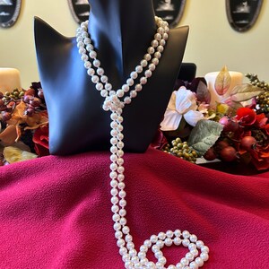 May include: A long white pearl necklace with a knotted design. The necklace is displayed on a black mannequin and draped over a red fabric surface.