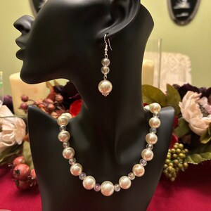 May include: A white pearl necklace and matching earrings. The necklace is made with large white pearls and smaller clear iridescent beads. The earrings are long and feature a single large white pearl at the bottom.