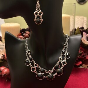 May include: A silver necklace with black oval-shaped stones and a matching pair of earrings. The necklace has a double chain design with a geometric pattern.
