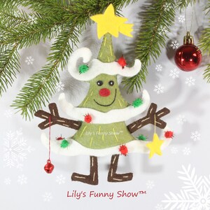 K&ouml;nnte beinhalten: Ein Weihnachtsbaum-Anh&auml;nger aus Filz mit einem l&auml;chelnden Gesicht, braunen Armen und Beinen. Der Baum ist mit wei&szlig;em Filz, gr&uuml;nen und roten Bommeln und einem gelben Stern oben verziert. Der Anh&auml;nger h&auml;ngt an einem wei&szlig;en Hintergrund mit Schneeflocken.