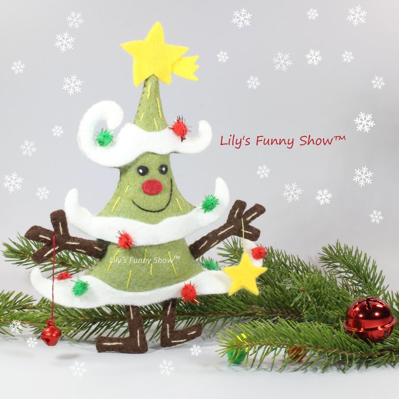 K&ouml;nnte beinhalten: Ein gr&uuml;ner Filz-Weihnachtsbaum mit wei&szlig;em Filzbesatz und einem gelben Stern oben. Der Baum hat ein l&auml;chelndes Gesicht und braune Filz-Arme und -Beine. Der Baum ist mit roten und gr&uuml;nen Filz-Ornamenten geschm&uuml;ckt. Der Baum steht auf einem gr&uuml;nen Kiefern-Zweig mit einem roten Glocken-Anh&auml;nger.