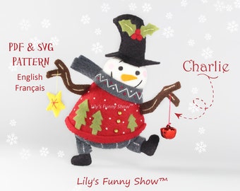Könnte beinhalten: Ein Filz-Schneemann-Anhänger namens Charlie, der einen roten Pullover mit grünen Bäumen und goldenen Knöpfen, einen grauen Schal und einen schwarzen Zylinder mit Stechpalme trägt. Er hält einen goldenen Stern in der einen Hand und eine rote Glocke in der anderen. Der Hintergrund ist weiß mit Schneeflocken.