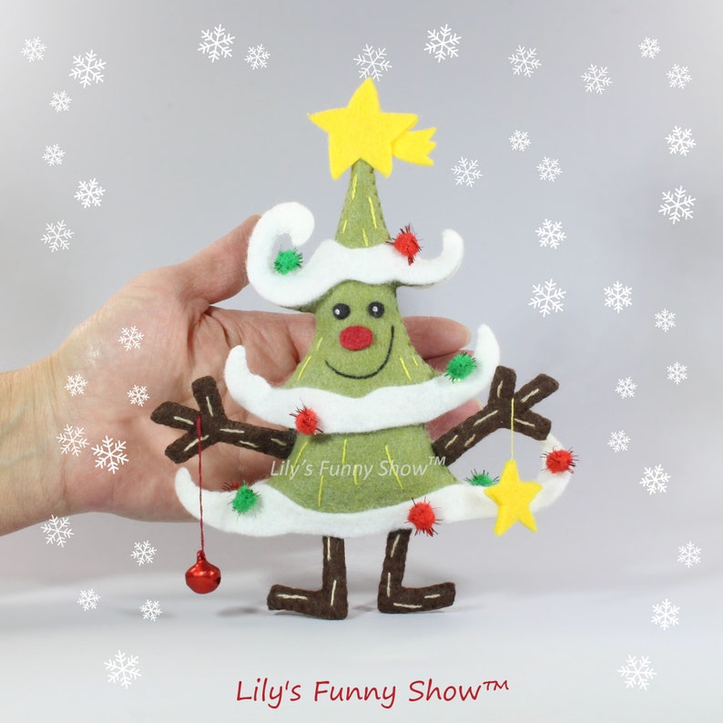 K&ouml;nnte beinhalten: Ein Filz-Weihnachtsbaum-Anh&auml;nger mit einem l&auml;chelnden Gesicht, wei&szlig;em Schnee, gr&uuml;nen Zweigen, braunen Beinen und einem gelben Stern oben. Der Anh&auml;nger h&auml;ngt an einer roten Kugel und einem gelben Stern. Der Text "Lily's Funny Show&trade;" befindet sich auf dem Anh&auml;nger.