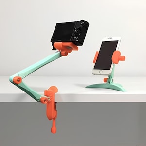 May include: Two phone and camera holders with adjustable arms. The holders are made of light blue and orange plastic. The holders are mounted on a white surface.