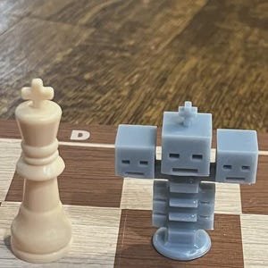 Peut inclure: Une pièce d'échecs en 3D imprimée en gris en forme de personnage Minecraft se tient sur un échiquier en bois. La pièce a une tête carrée avec une croix sur le dessus et un corps fait de blocs empilés. Une pièce de roi d'échecs blanche est en arrière-plan.