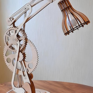 May include: A wooden desk lamp with a gear-shaped base and a slatted shade. The lamp is made of light brown wood and has a white cord.