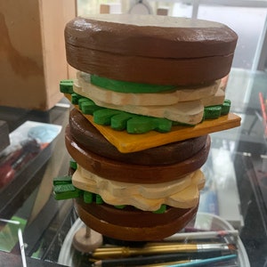 May include: A wooden toy hamburger with brown buns, green lettuce, yellow cheese, and white cheese slices.