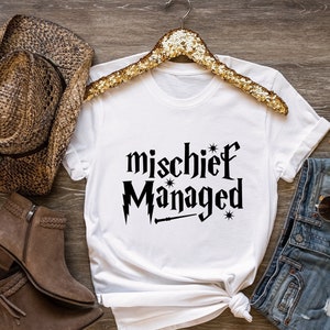 May include: White t-shirt with black text that reads "mischief managed" with a wand and stars. The shirt is styled with a brown cowboy hat, brown boots, and denim shorts.