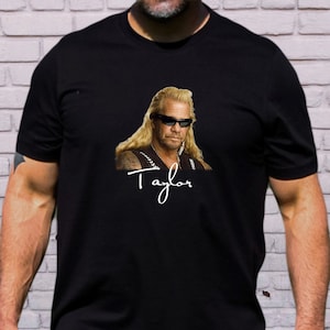 May include: Black t-shirt featuring a color image of a man with long blonde hair and sunglasses. The man is wearing a leather jacket and has a signature that says "Taylor" underneath the image.