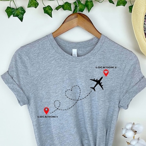 Puede incluir: Camiseta gris con un avi&oacute;n negro volando sobre una l&iacute;nea en forma de coraz&oacute;n. La l&iacute;nea conecta dos alfileres rojos etiquetados como "LOCATION 1" y "LOCATION 2".
